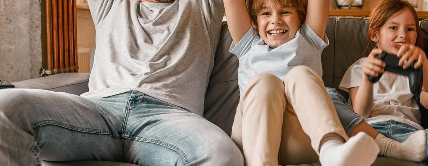 Eine junge Familie mit zwei Kindern sitzt auf einem Sofa und hält Consolen in der Hand. Im Hintergrund ist eine Küchenzeile und angeschaltete Lampen.