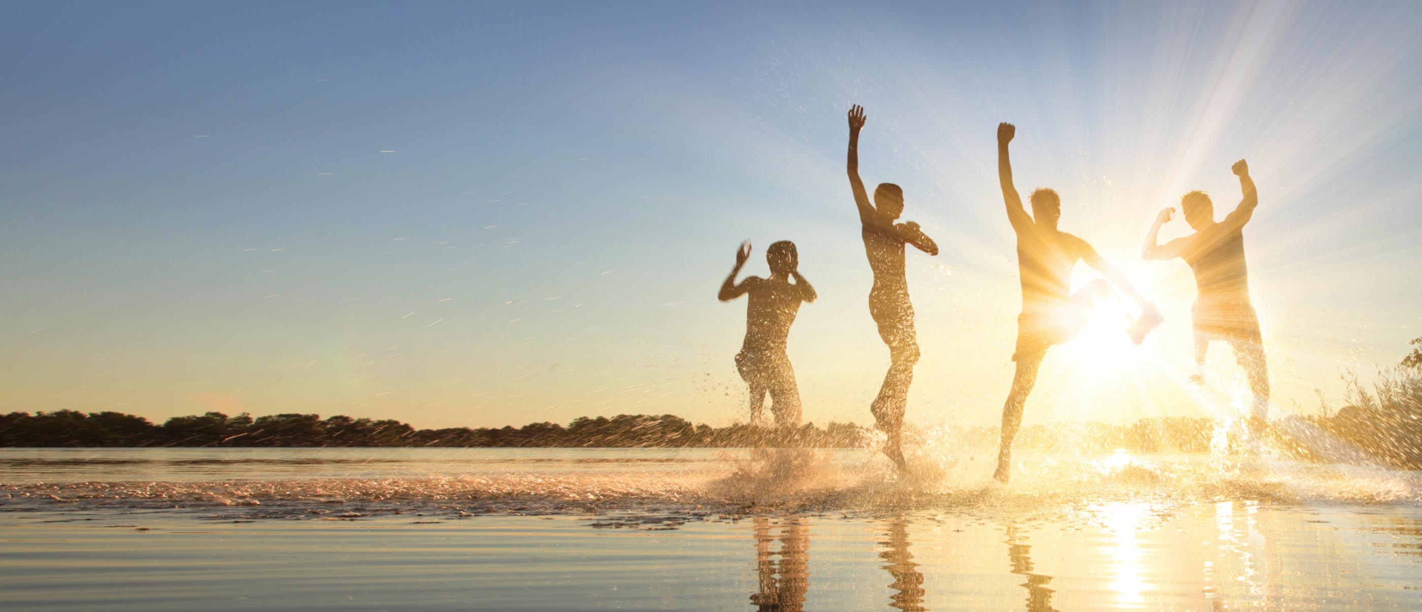 Menschen Springen am Strand in die Höhe. Die Sonne scheint im Hintergrund.